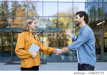 Two smiling business people shaking hands in front of a modern glass building, signifying a successful agreement, teamwork, and a new professional relationship Two smiling business people shaking hands in front of a modern glass building, signifying a successful agreement, teamwork, and a new professional relationship 138835735