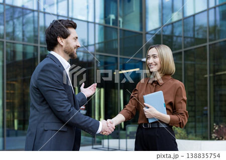 Two smiling business people warmly shaking hands outside a modern office building, signifying a successful partnership, agreement, professionalism, and teamwork in a corporate environment Two smiling business people warmly shaking hands outside a modern office building, signifying a successful partnership, agreement, professionalism, and teamwork in a corporate environment 138835754