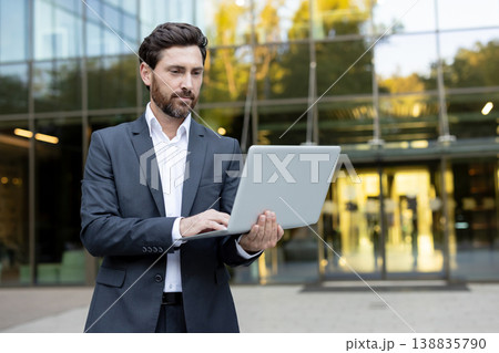 Confident businessman standing outside a contemporary glass building, focused on typing and working on a laptop, symbolizing outdoor work, remote collaboration, and business lifestyle Confident businessman standing outside a contemporary glass building, focused on typing and working on a laptop, symbolizing outdoor work, remote collaboration, and business lifestyle 138835790