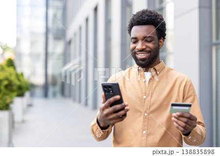 Smiling young person holding a smartphone and credit card, performing an online payment or banking transaction outdoors, representing digital commerce and mobile financial services Smiling young person holding a smartphone and credit card, performing an online payment or banking transaction outdoors, representing digital commerce and mobile financial services 138835898