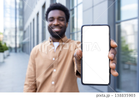 Smiling man holding smartphone vertically with blank white screen for mockup, standing outdoors by a modern office building, showcasing mobile app, advertising and digital display concepts 138835903