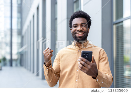 Happy young man celebrating a victory, holding a mobile phone and making a fist of joy, standing outdoors in front of a modern building, representing achievement and business success Happy young man celebrating a victory, holding a mobile phone and making a fist of joy, standing outdoors in front of a modern building, representing achievement and business success 138835909