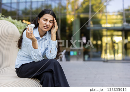 Young business woman sitting on a modern bench outdoors, touching her temple in pain, holding a tissue, depicting stress, burnout, and migraine while experiencing a significant headache 138835913