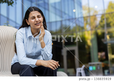 Young indian woman grimacing in pain, holding her cheek on a city bench, suffering from intense toothache and jaw discomfort while dressed in business casual outdoors 138835919