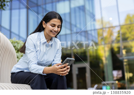Young businesswoman smiling, sitting outdoors and using a mobile phone, representing remote work, digital communication, and modern technology in a corporate environment Young businesswoman smiling, sitting outdoors and using a mobile phone, representing remote work, digital communication, and modern technology in a corporate environment 138835920