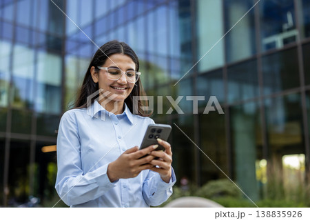 Young professional woman smiling happily while interacting with her smartphone, checking messages, browsing, or working on a mobile application in a modern urban environment Young professional woman smiling happily while interacting with her smartphone, checking messages, browsing, or working on a mobile application in a modern urban environment 138835926