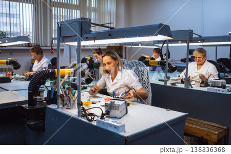 Women at their desks solder radio components to electronic boards. Factory for the production of electronic equipment. Women at their desks solder radio components to electronic boards. Factory for the production of electronic equipment. 138836368