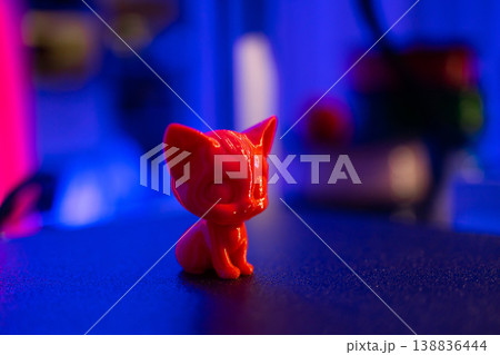 A small, glossy red cat figurine sits on a dark surface illuminated by vibrant neon blue and pink lights, creating a moody atmosphere A small, glossy red cat figurine sits on a dark surface illuminated by vibrant neon blue and pink lights, creating a moody atmosphere 138836444
