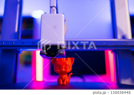 A 3D printer is in the process of creating a small, bright orange cat figurine with intricate details 138836448