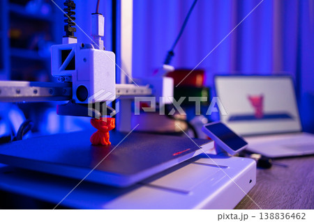A 3D printer is actively creating a small orange object on its build plate, with a laptop displaying the design in the background A 3D printer is actively creating a small orange object on its build plate, with a laptop displaying the design in the background 138836462