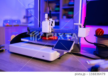 A modern 3D printer is actively creating a red plastic object on its build plate with filament spool nearby 138836479