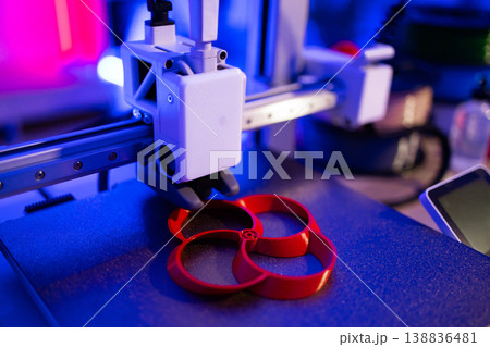 A 3D printer is actively creating a red, circular object on its build plate under vibrant blue and pink lighting 138836481