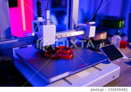 A modern 3D printer is actively creating a red object layer by layer on its build plate with vibrant blue and pink lighting 138836482