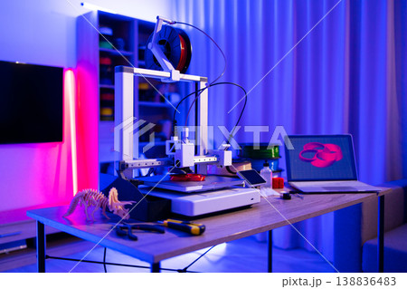 A 3D printer is actively creating a red object on its platform, with filament spools and a laptop displaying a 3D model nearby 138836483