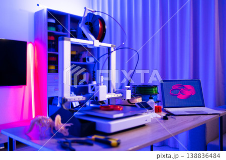 A 3D printer is actively creating an object while a laptop displays its design, surrounded by filament spools and tools under vibrant neon lighting A 3D printer is actively creating an object while a laptop displays its design, surrounded by filament spools and tools under vibrant neon lighting 138836484