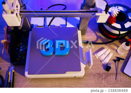 A 3D printer is shown with spools of filament, tools, and other accessories laid out on a wooden surface under blue and purple lighting A 3D printer is shown with spools of filament, tools, and other accessories laid out on a wooden surface under blue and purple lighting 138836489