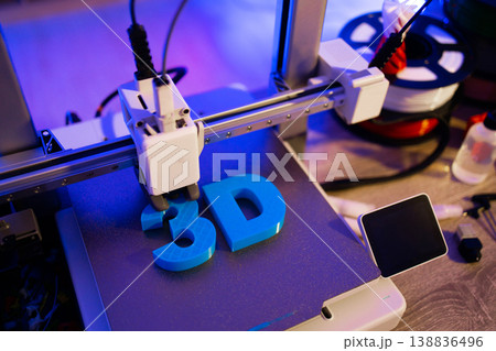 A 3D printer is actively creating blue letters on its build plate with filament spools in the background 138836496