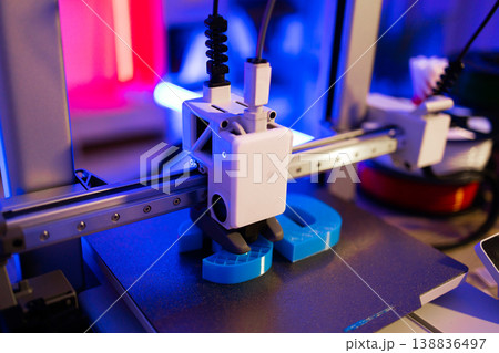A modern 3D printer is actively creating a blue object layer by layer with vibrant neon lighting 138836497