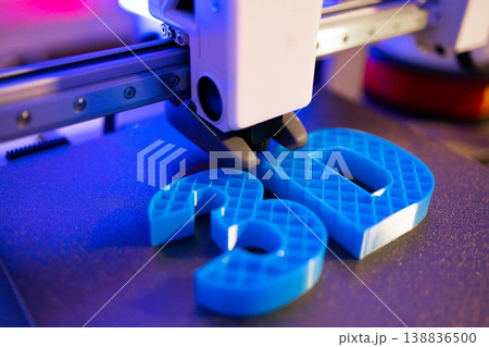 A close-up shot shows a 3D printer's nozzle depositing blue plastic to form the numbers 3 and 0 on a textured surface under purple lighting 138836500