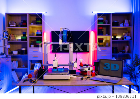 A modern 3D printer is actively creating an object on a desk, illuminated by vibrant neon lights, with a laptop displaying 3D graphics nearby A modern 3D printer is actively creating an object on a desk, illuminated by vibrant neon lights, with a laptop displaying 3D graphics nearby 138836511