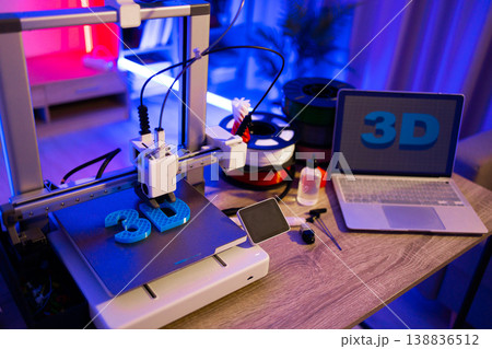 A 3D printer is actively creating blue plastic letters on its build plate, with spools of filament and a laptop displaying the 3D logo nearby 138836512