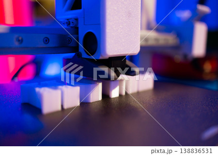 A close-up shot of a 3D printer's nozzle depositing material to build small white objects layer by layer with vibrant blue and pink lighting A close-up shot of a 3D printer's nozzle depositing material to build small white objects layer by layer with vibrant blue and pink lighting 138836531