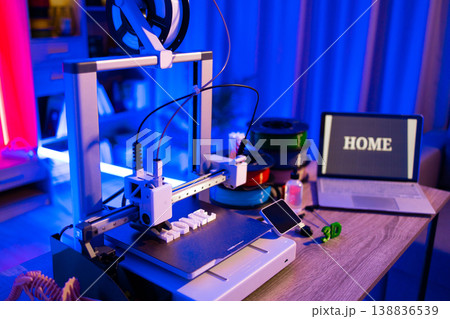 A 3D printer is actively creating the word HOME in white filament on its build plate, illuminated by blue and pink lights, with a laptop displaying the same word in the background 138836539