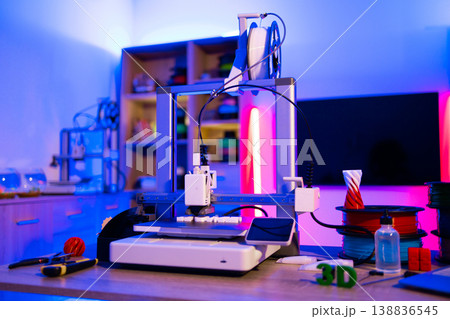 A modern 3D printer is set up in a room with vibrant blue and pink neon lighting, surrounded by spools of filament and tools 138836545