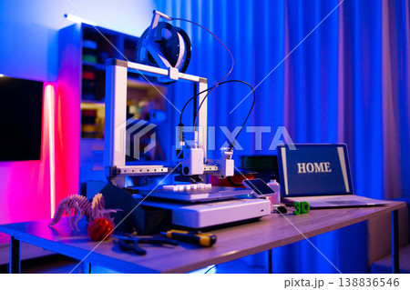 A 3D printer is actively creating a small object on its platform, surrounded by tools and a laptop displaying the word HOME 138836546