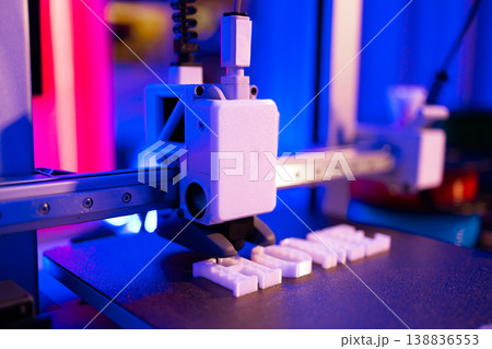 A 3D printer is actively creating white plastic letters on its build plate under vibrant blue and pink lighting A 3D printer is actively creating white plastic letters on its build plate under vibrant blue and pink lighting 138836553