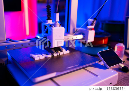 A modern 3D printer is actively creating white plastic letters on its build plate with vibrant neon lighting 138836554