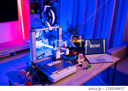 A 3D printer is actively creating the word HOME in white filament on its build plate, surrounded by spools of filament and a laptop displaying the same word 138836657