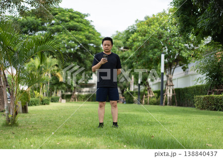 Asian man wearing black sportswear stands on grass in a residential park using a smartphone during an outdoor fitness activity. Asian man wearing black sportswear stands on grass in a residential park using a smartphone during an outdoor fitness activity. 138840437