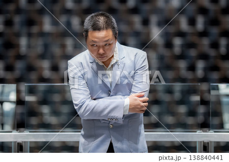 Formal portrait of an Asian businessman in pale suit leaning forward with folded arms indoors, with serious upward gaze and a soft bokeh background that keeps focus on his expression. 138840441