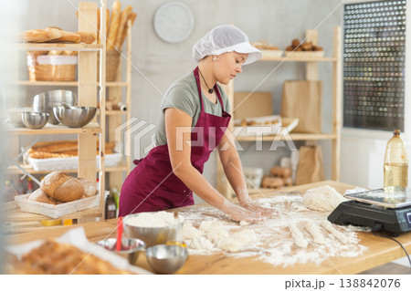 Female baker prepares raw dough in bakery, forming it into long sausages to be cut into pieces for baking buns or croissants Female baker prepares raw dough in bakery, forming it into long sausages to be cut into pieces for baking buns or croissants 138842076