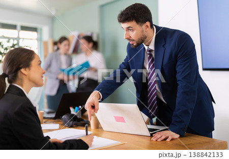 Male manager leaning over table and instructing young employee Male manager leaning over table and instructing young employee 138842133