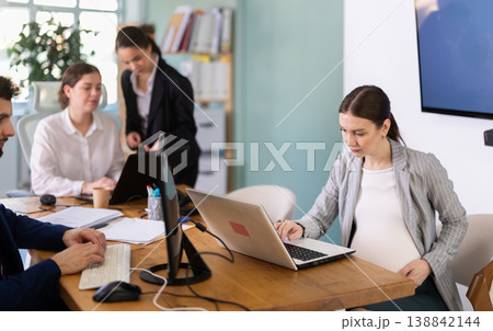 Pregnant woman is working on a laptop in an office while colleagues are discussing a project Pregnant woman is working on a laptop in an office while colleagues are discussing a project 138842144