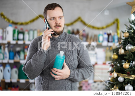 Handsome man speaking mobile and choose something in cosmetics store decorated for New Year holiday Handsome man speaking mobile and choose something in cosmetics store decorated for New Year holiday 138842205