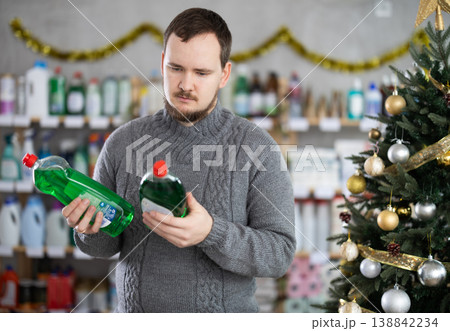 Focused young man choosing dishwashing liquid on supermarket shelf decorated for New Year holiday Focused young man choosing dishwashing liquid on supermarket shelf decorated for New Year holiday 138842234