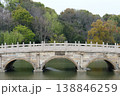 Stone Arch Bridge Over Calm Park Lake Surrounded by Lush Trees and Autumn Foliage 138846259