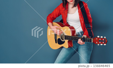 Female musician in red jacket and jeans playing an acoustic guitar against blue studio background Female musician in red jacket and jeans playing an acoustic guitar against blue studio background 138846465