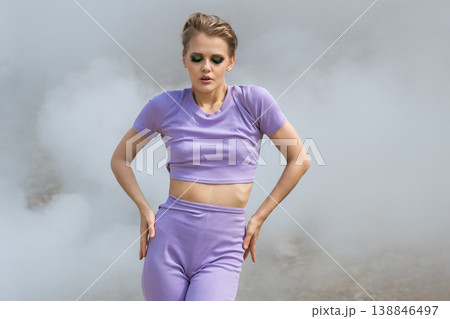 Stylish woman in lavender activewear with bold green eyeshadow posing in dramatic fog Stylish woman in lavender activewear with bold green eyeshadow posing in dramatic fog 138846497