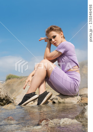 Woman with short hair in a lavender outfit posing on rocks by a natural hot spring under a blue sky Woman with short hair in a lavender outfit posing on rocks by a natural hot spring under a blue sky 138846498