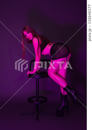 Young woman dressed in lingerie and stripper platform heels stands leaning on bar stool 138846577