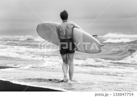 Woman from behind holding surfboard walking on shore looking out at ocean waves 138847754
