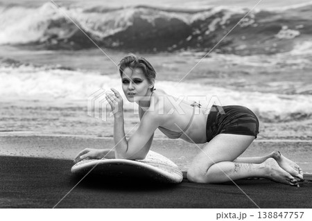 Monochrome fashion portrait of young woman with surfboard kneeling on dark sand beach Monochrome fashion portrait of young woman with surfboard kneeling on dark sand beach 138847757