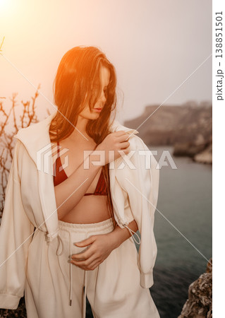 Woman coast hoodie posing on a rocky shore wearing a white tracksuit and bikini top during a golden sunset at the sea Woman coast hoodie posing on a rocky shore wearing a white tracksuit and bikini top during a golden sunset at the sea 138851501