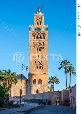 Kutubiyya Mosque is visible in Marrakesh. The mosque features a tall minaret surrounded by palm trees. People walk nearby, enjoying the sunny day in a lively area of the city. 138852247