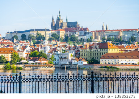 The scene shows Prague Castle rising above the city. Houses and trees line the river below. Many buildings have red roofs. The clear sky makes for a bright day in Prague. The scene shows Prague Castle rising above the city. Houses and trees line the river below. Many buildings have red roofs. The clear sky makes for a bright day in Prague. 138852275