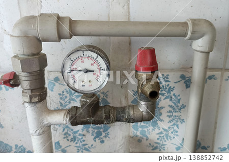 Close-up of a household plumbing system with pressure gauge, safety valve, and pipes mounted on a tiled wall, showing water pressure control equipment in a domestic setting 138852742
